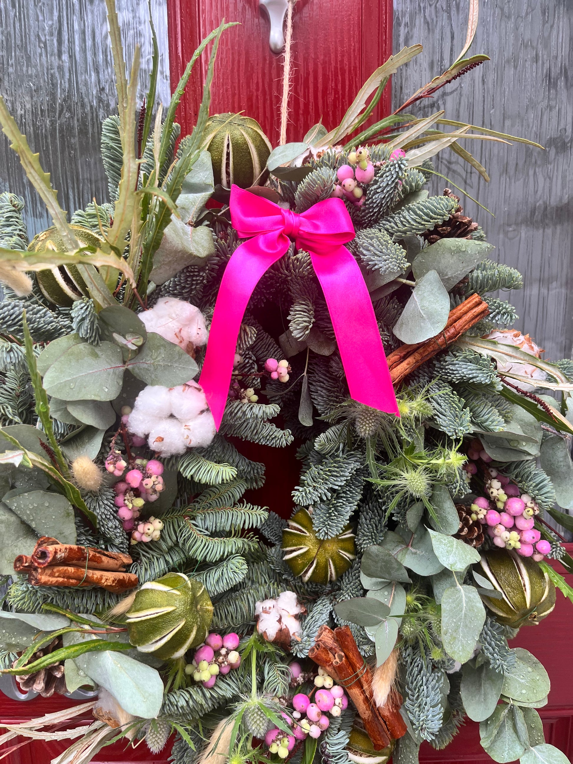 Decorative wreath with greenery, pink ribbon, and cinnamon sticks against a red door.
