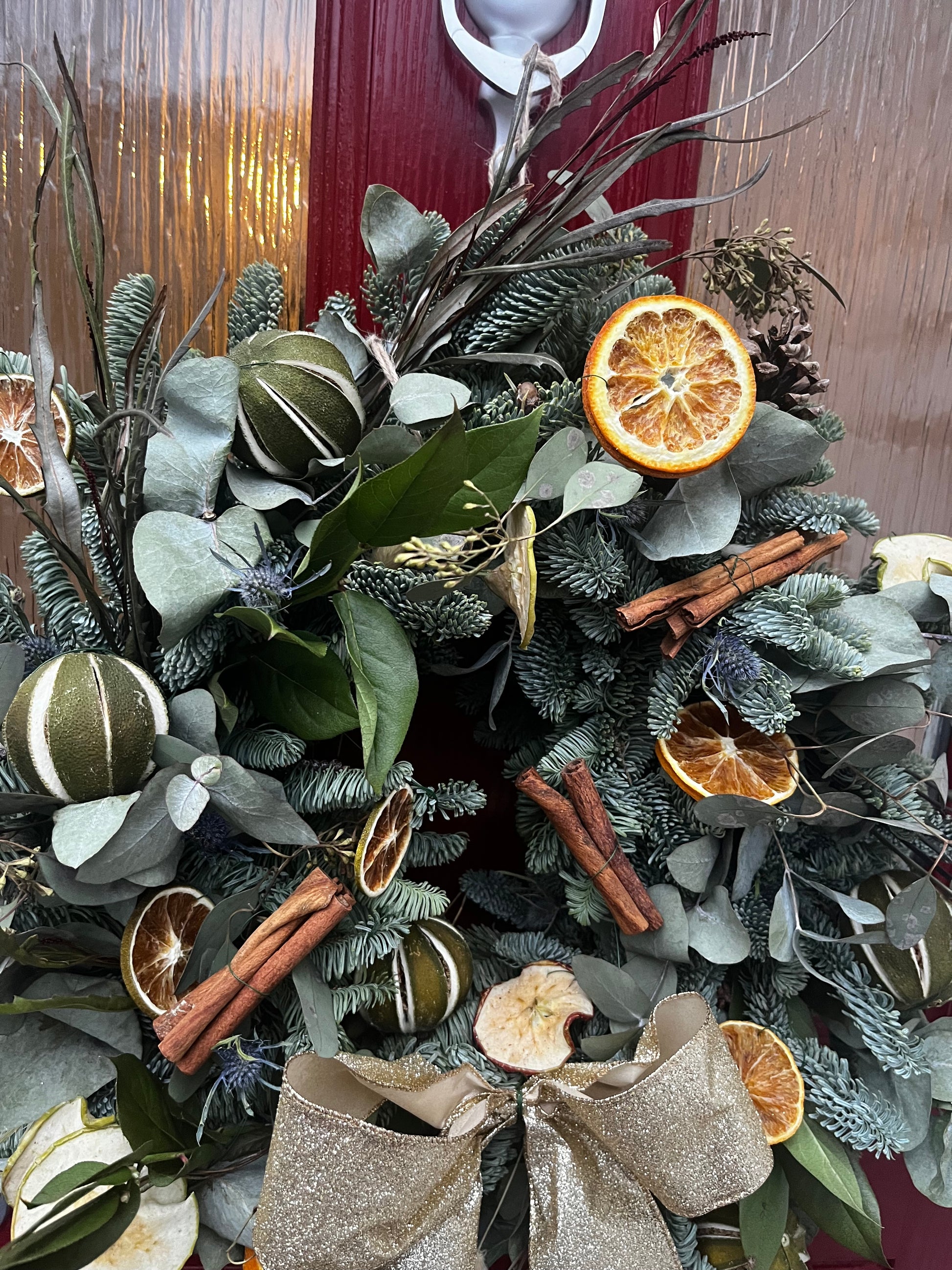 Decorative wreath with greenery, oranges, cinnamon sticks, and a bow on a wooden surface.