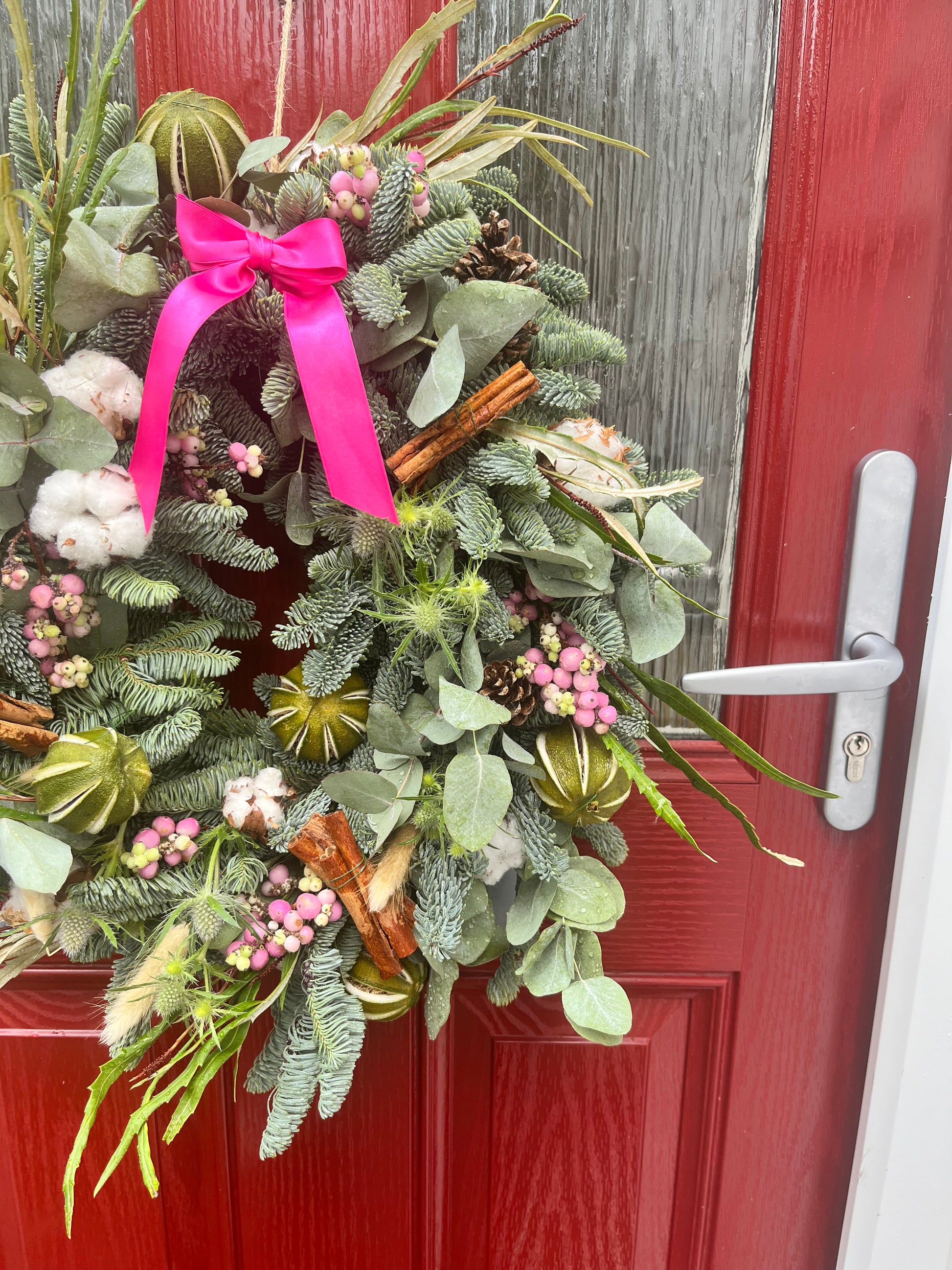 Decorative wreath with greenery, pink ribbon, and cinnamon sticks on a red door.