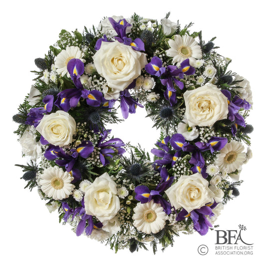 A stunning wreath featuring white roses, iris, thistle, and seasonal greenery, beautifully crafted in Stevenage to offer a peaceful and elegant tribute.